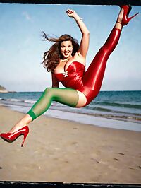 A statuesque brunette woman in a red backless dress shines brightly on a twilight beach