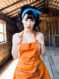 A tousled haired apron clad woman in 1950s style stands in a dusty barn at high noon